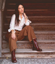 Wearing the Isodora handcrafted Italian leather boots by Bed|Stü, a woman in a white blouse and brown wide-leg pants sits on worn wooden stairs, gazing off to the side with one hand resting on her neck.
