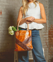 A woman in a sleeveless white top and jeans holds the Bed|Stü Italic brown leather tote bag, featuring padded shoulder straps and an organic cotton lining, with flowers peeking out from inside as she stands against a brick wall.