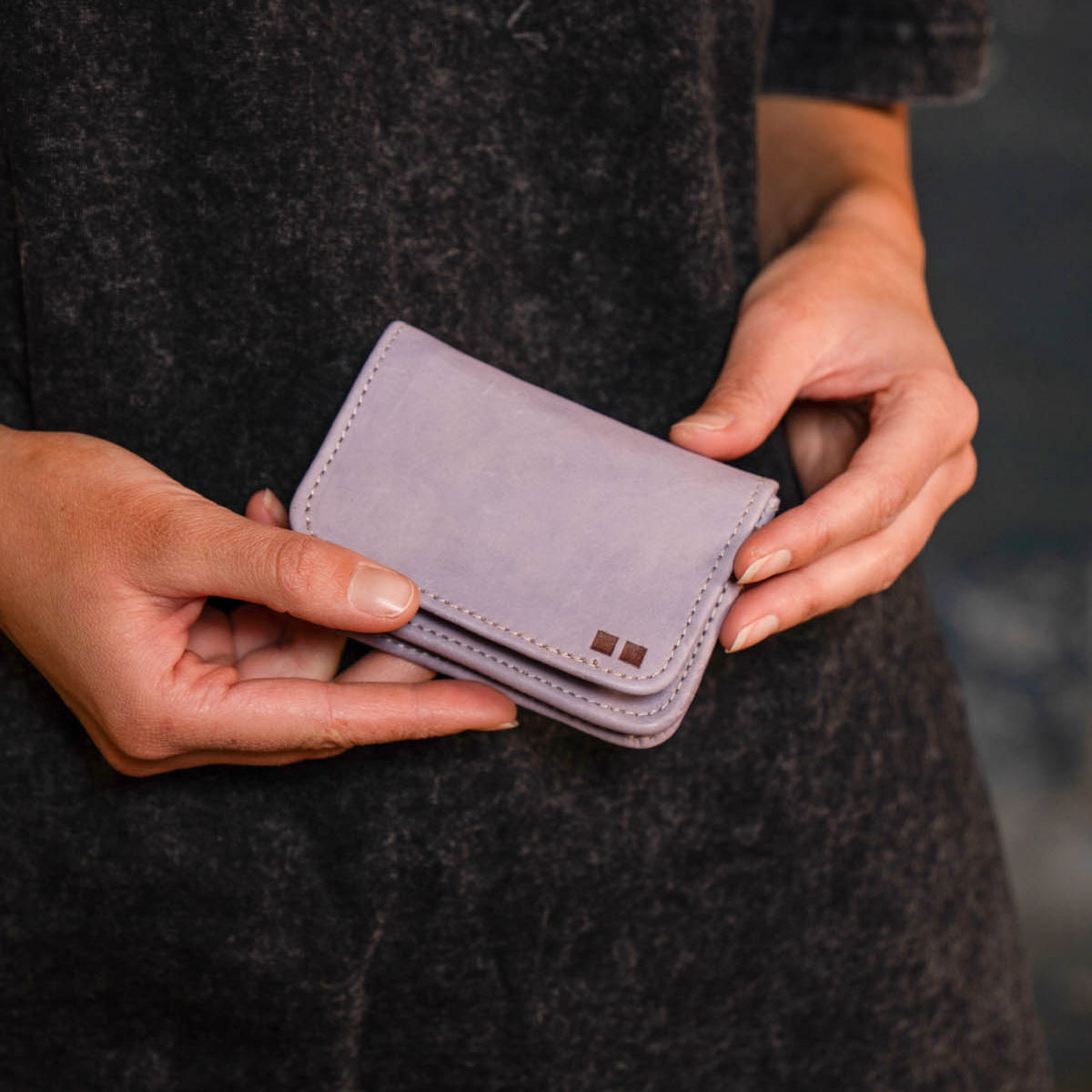 A person holds the Bed|Stü Jeor, a small, light purple compact wallet, with both hands against a dark textured background.