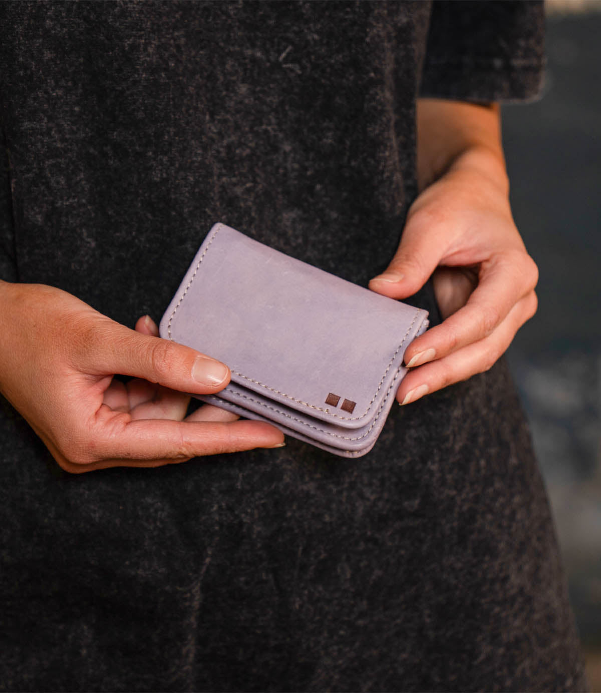 A person holds the Bed|Stü Jeor, a small, light purple compact wallet, with both hands against a dark textured background.