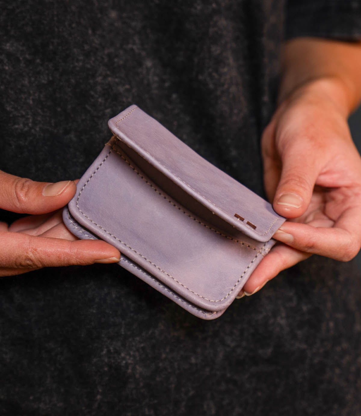 A person holds the Bed|Stü Jeor compact wallet—a small, light purple accessory—with both hands against a dark background.