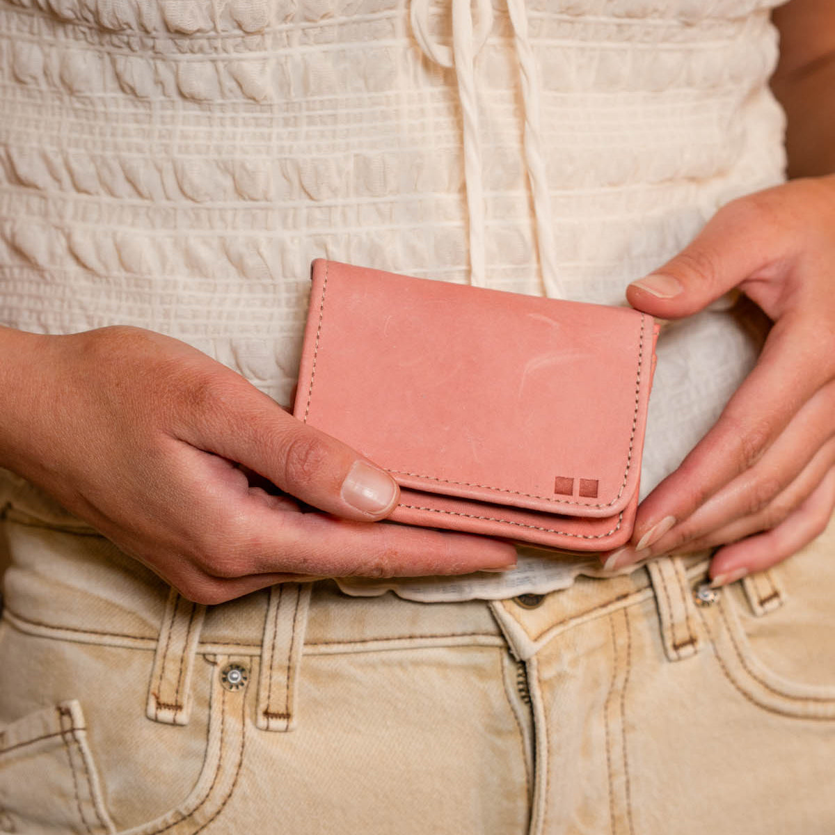 A person in a white top and beige pants holds the Bed|Stü Jeor compact wallet with both hands in front of their waist.