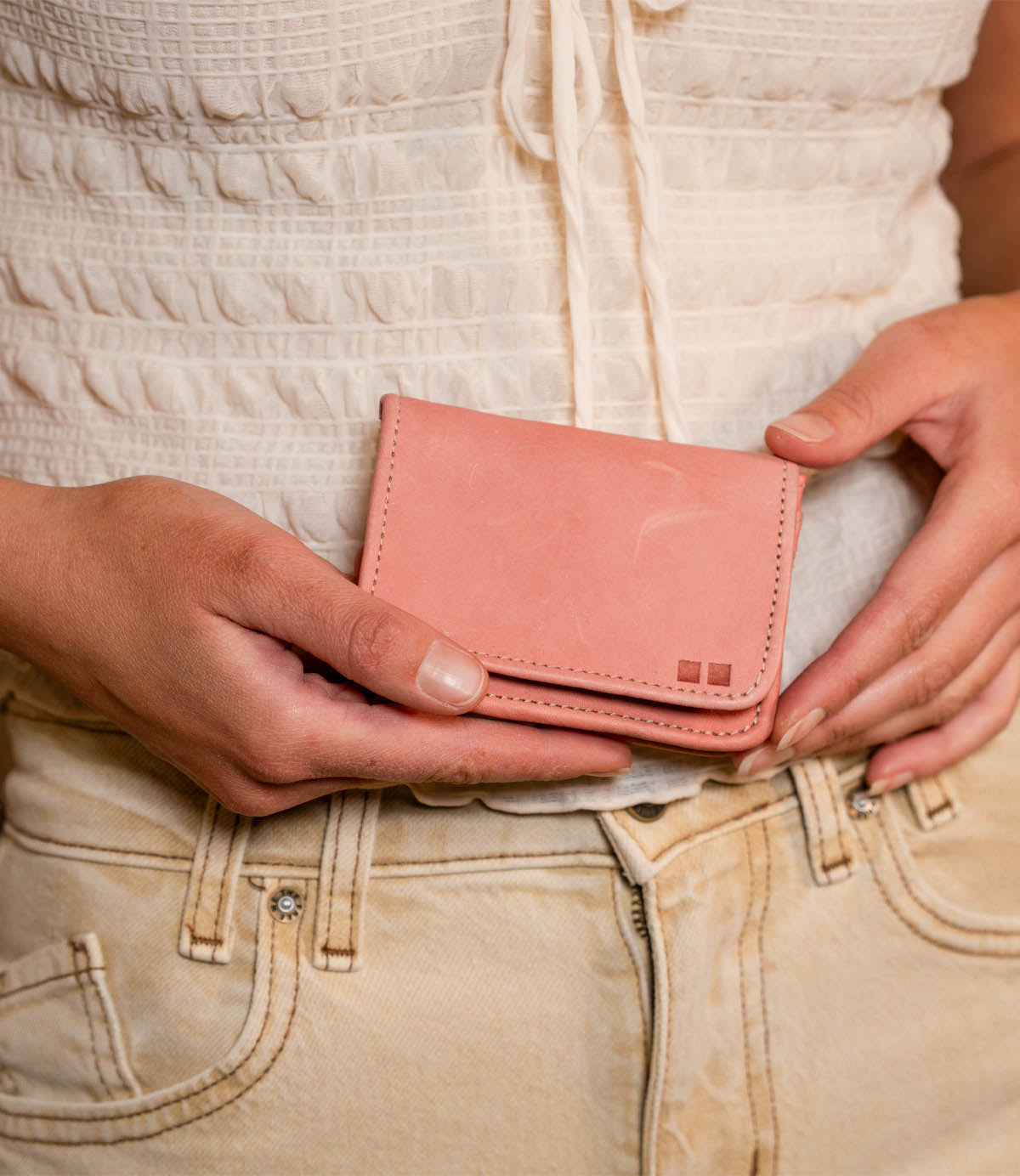A person in a white top and beige pants holds the Bed|Stü Jeor compact wallet with both hands in front of their waist.