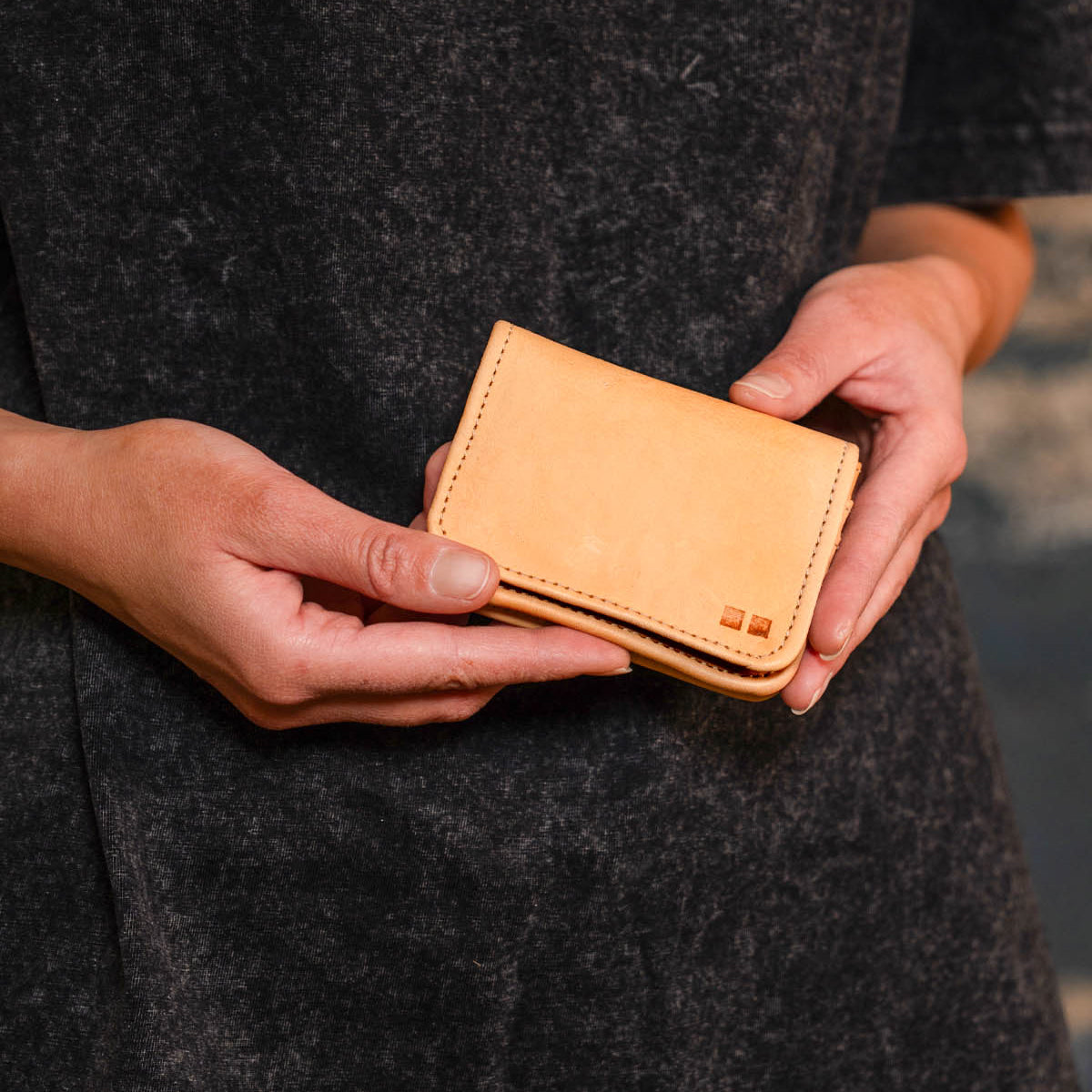 A person wearing a dark garment holds the Jeor compact card wallet by Bed|Stü with both hands against a neutral background. This small, rustic leather accessory features convenient credit card slots, seamlessly blending style and practicality in an understated design.
