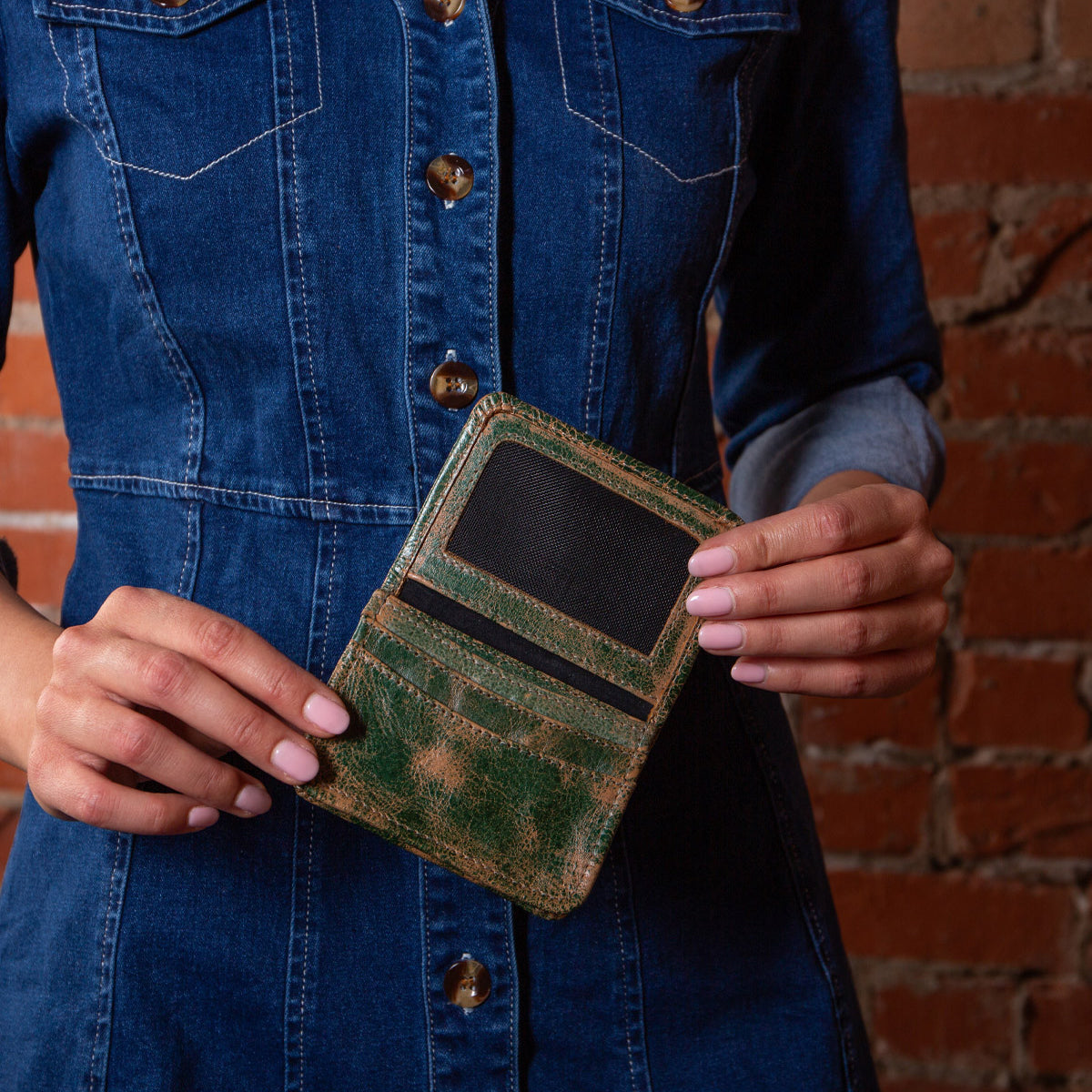 A person in a denim dress holds the open Bed|Stü Jeor compact wallet, showing its credit card slots against a brick wall background.