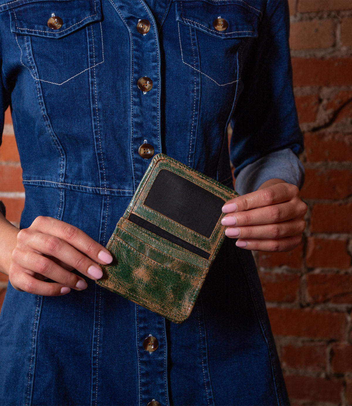 A person in a denim dress holds the open Bed|Stü Jeor compact wallet, showing its credit card slots against a brick wall background.
