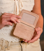 A person in a white top and beige pants holds the Bed|Stü Jeor wallet open, revealing its rustic leather design with credit card slots and a transparent ID window.