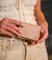 A person wearing a white blouse and beige pants holds the Jeor wallet by Bed|Stü with both hands. This compact, small card wallet is crafted from rustic light brown leather and features convenient credit card slots, providing both style and functionality.