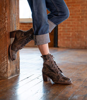 A person in cuffed jeans leans against a wooden beam, wearing the Bed|Stü Judgement brown leather lace-up boots with a cap toe and low block heel on a polished wooden floor in a brick-walled room.