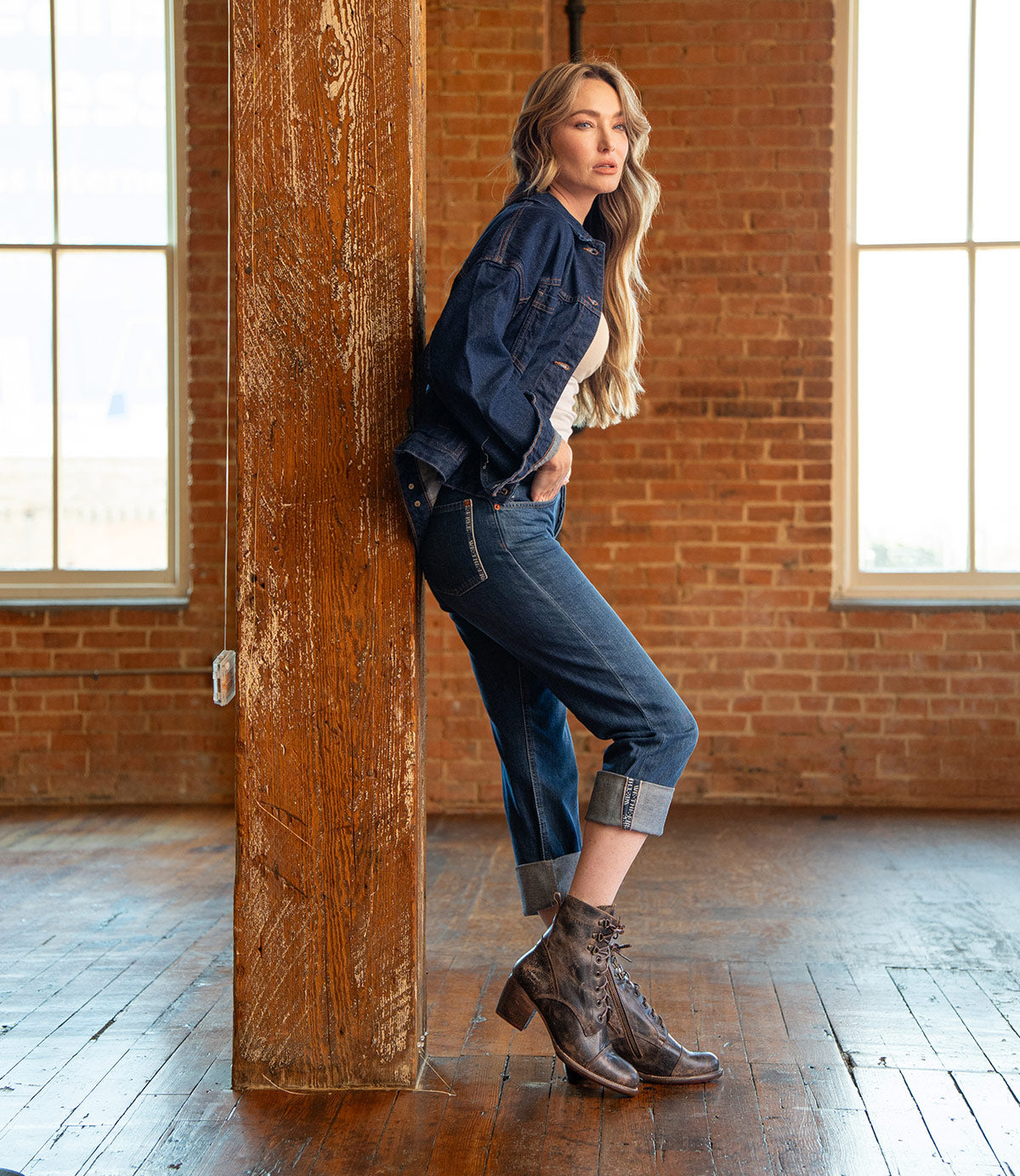 A woman in a denim jacket and cuffed jeans leans against a wooden beam in a brick-walled room with large windows, wearing the stylish cap-toe Judgement boots by Bed|Stü.