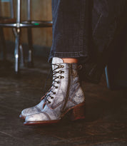 A person wearing Bed|Stü Judgement metallic silver leather lace-up ankle boots with side zippers, cap toe, and sturdy block heels, paired with dark cuffed pants. The Judgement boots rest on a dark wooden floor next to a metal chair.