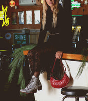 A woman in a black lace dress sits on a bar counter, holding a red purse and wearing Bed|Stü Judgement gray leather lace-up boots with a stylish block heel. Pine branches are placed on the counter.