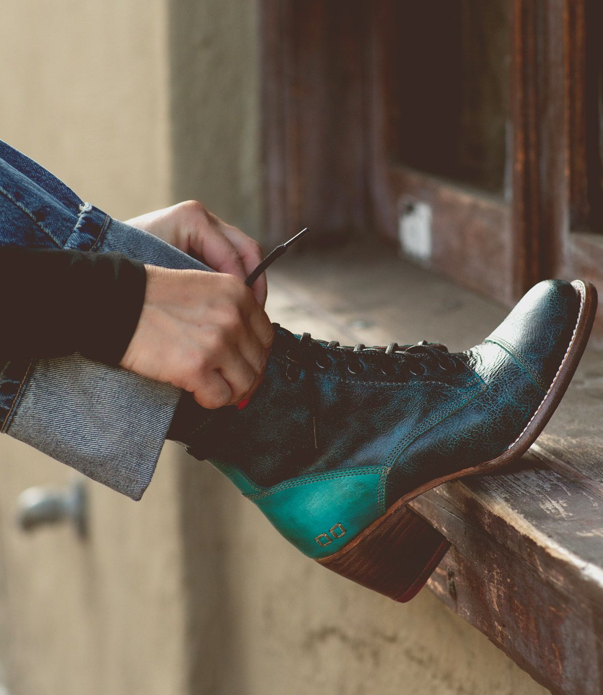 A person sits on a windowsill in blue jeans, tying the laces of the Bed|Stü Judgement boot—a teal and black leather lace-up with a sleek cap toe and wooden heel.
