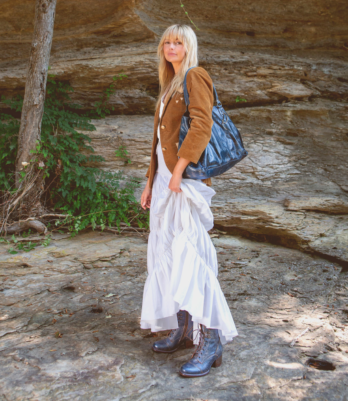 A woman with blonde hair stands outdoors on rocky ground, wearing a brown jacket, a white dress, and the Bed|Stü Judgement striking blue leather lace-up boots featuring a block heel and cap toe. She holds a large black bag, with green plants and a rock wall in the background.