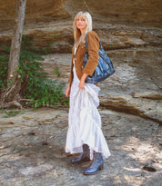 A woman with blonde hair stands outdoors on rocky ground, wearing a brown jacket, a white dress, and the Bed|Stü Judgement striking blue leather lace-up boots featuring a block heel and cap toe. She holds a large black bag, with green plants and a rock wall in the background.