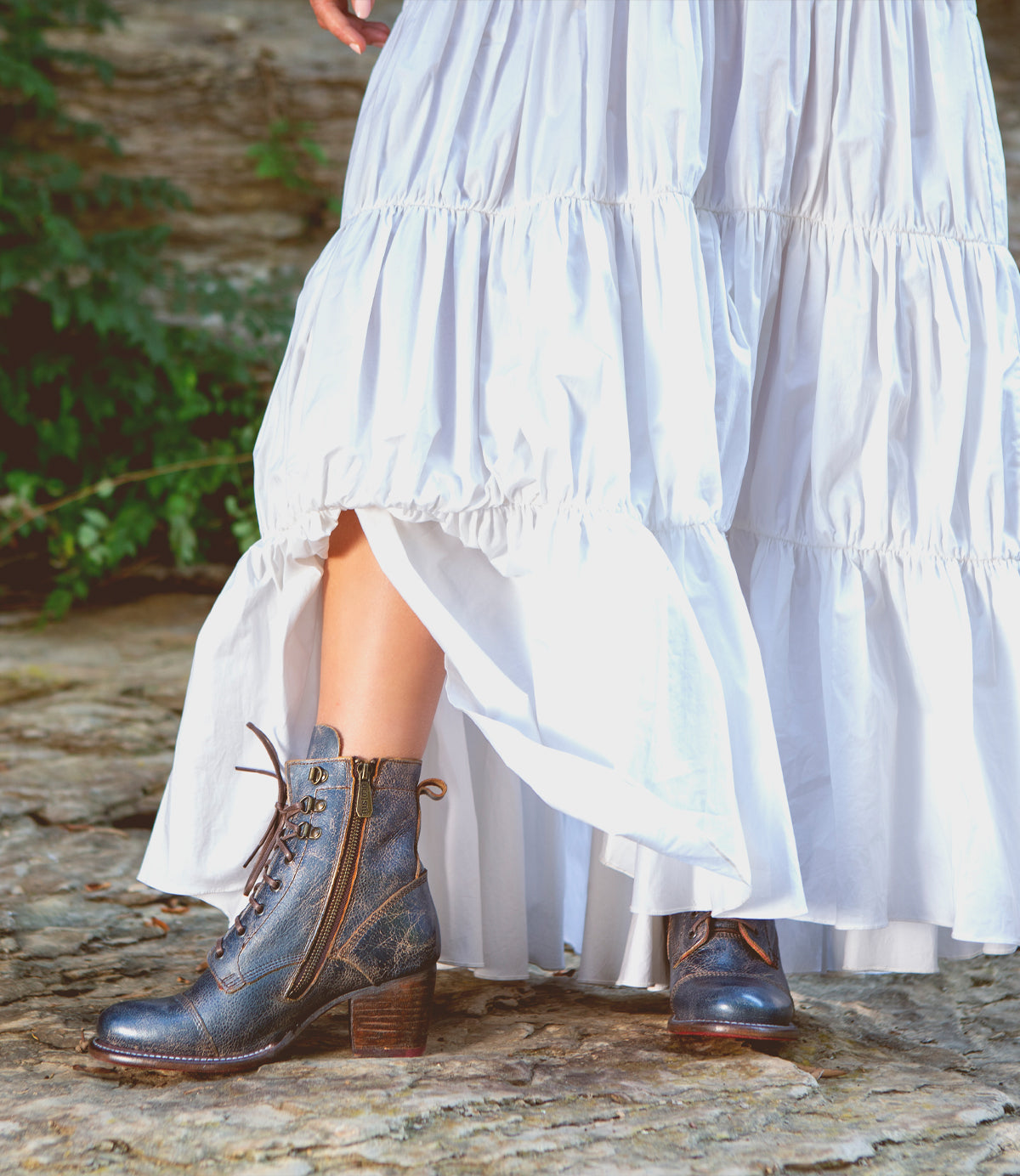 A person wearing a long, white, ruffled dress and Bed|Stü Judgement blue leather lace-up ankle boots with cap toe, zippers, and a stylish block heel, standing on a stone surface outdoors.