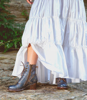 A person wearing a long, white, ruffled dress and Bed|Stü Judgement blue leather lace-up ankle boots with cap toe, zippers, and a stylish block heel, standing on a stone surface outdoors.