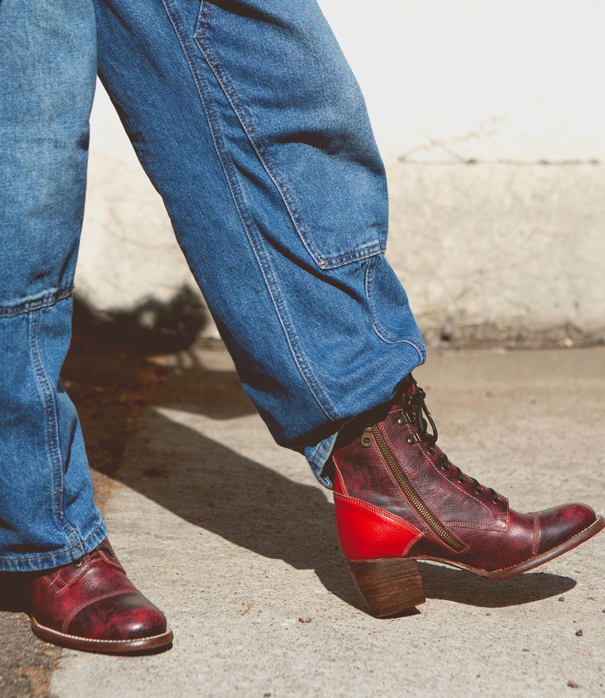 A person wearing blue jeans and a pair of Bed|Stü Judgement red leather lace-up boots with a low block heel and classic cap toe, standing on a sunlit concrete surface.