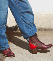 A person wearing blue jeans and a pair of Bed|Stü Judgement red leather lace-up boots with a low block heel and classic cap toe, standing on a sunlit concrete surface.