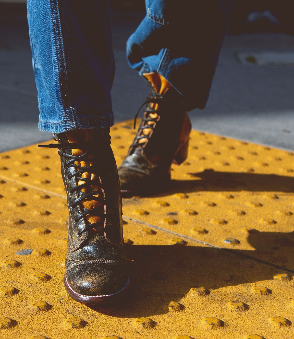 A person wearing Bed|Stü Judgement black cap toe leather lace-up boots and blue jeans stands on a yellow textured sidewalk surface, likely a tactile paving tile for visually impaired pedestrians.