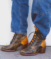 A person wears Bed|Stü's Judgement faded blue jeans and distressed brown leather lace-up boots with a cap toe, standing on a gray concrete surface in front of a white background.