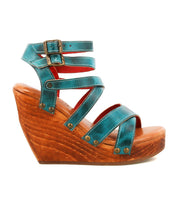 The Juliana II by Bed|Stü is a turquoise strappy wedge sandal with a wooden platform, featuring a strappy upper, two buckles at the ankle, and metallic rivet details on the straps.