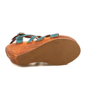 The Juliana II by Bed|Stü is shown from the bottom, featuring its solid sole and the textured wood grain platform beneath a strappy teal upper.
