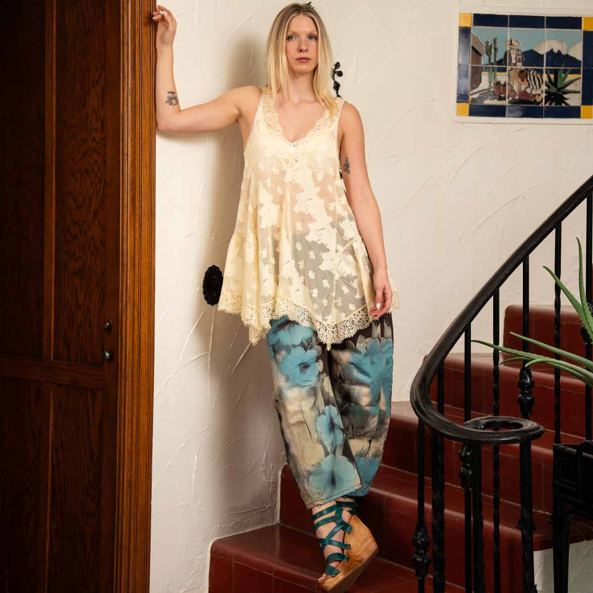 A woman in a loose cream lace top and blue tie-dye pants stands on red-tiled stairs next to a wooden door and a wall adorned with framed art, her look completed by the chic Bed|Stü Juliana II platform sandals featuring a stylish strappy upper.