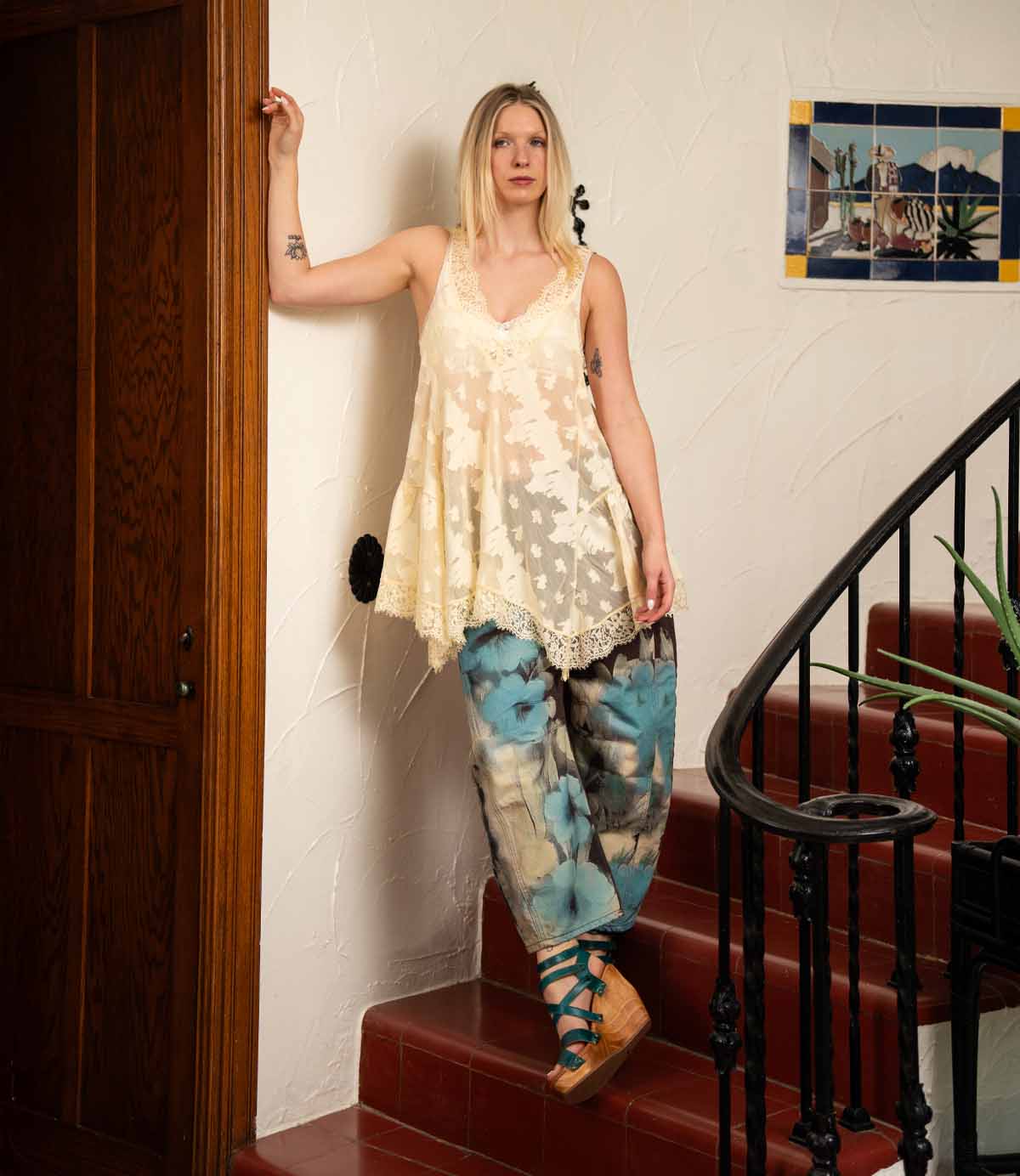 A woman in a loose cream lace top and blue tie-dye pants stands on red-tiled stairs next to a wooden door and a wall adorned with framed art, her look completed by the chic Bed|Stü Juliana II platform sandals featuring a stylish strappy upper.