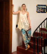 A woman in a loose cream lace top and blue tie-dye pants stands on red-tiled stairs next to a wooden door and a wall adorned with framed art, her look completed by the chic Bed|Stü Juliana II platform sandals featuring a stylish strappy upper.