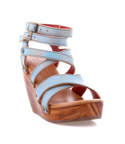 The Juliana II wedge sandal by Bed|Stü is shown from the front on a white background, featuring a light blue strappy upper, ankle straps with buckle closures, and a wooden platform wedge sole.