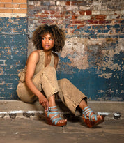 A woman with curly hair sits on a concrete floor against a weathered, painted brick wall, wearing tan clothes and blue Bed|Stü Juliana II platform sandals featuring a stylish strappy upper.