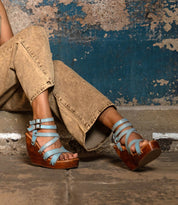 A person wearing tan jeans and the Juliana II blue strappy upper wedge sandals by Bed|Stü sits on a rough concrete floor against a textured blue and brown wall.