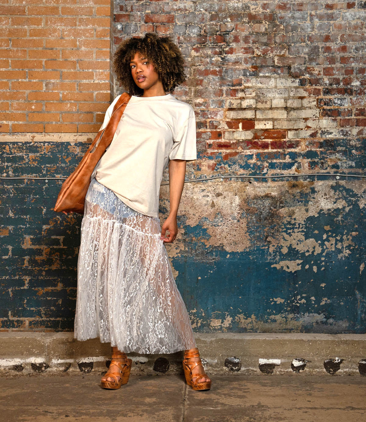 A woman stands against a worn brick wall, wearing a beige t-shirt, sheer lace skirt, and a brown shoulder bag. She completes her look with the Bed|Stü Juliana II platform sandals, featuring a strappy upper and wooden wedge.