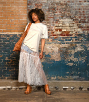A woman stands against a worn brick wall, wearing a beige t-shirt, sheer lace skirt, and a brown shoulder bag. She completes her look with the Bed|Stü Juliana II platform sandals, featuring a strappy upper and wooden wedge.
