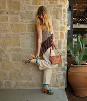 A woman in beige pants and a striped top adjusts her Bed|Stü Kalah teal platform heel with an adjustable ankle strap while standing by a stone wall adorned with potted cacti; she holds a brown bag and looks down at her foot.