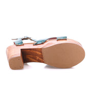 The Bed|Stü Kalah sandal—a single women's shoe featuring green straps, visible rivets, a sustainably sourced wooden platform sole, and a platform heel—is shown lying on its side to display the underside.