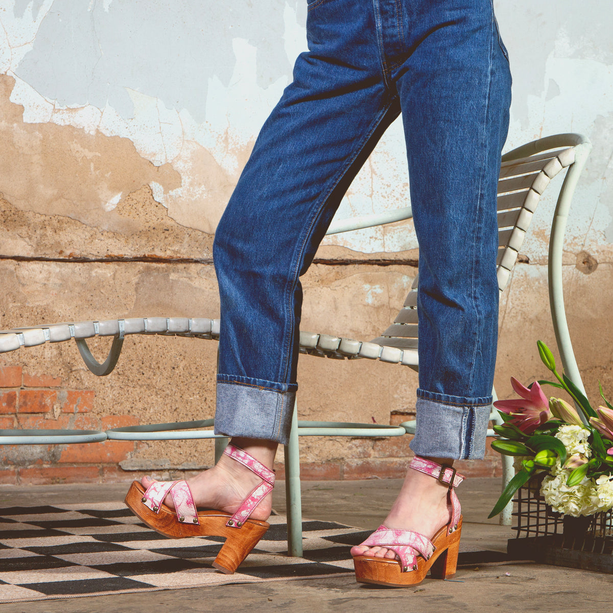 Wearing the Kalah sandals by Bed|Stü—featuring a pink platform design with an adjustable ankle strap—a person in cuffed jeans stands on a patterned tile floor near a white chair and a bouquet of flowers.