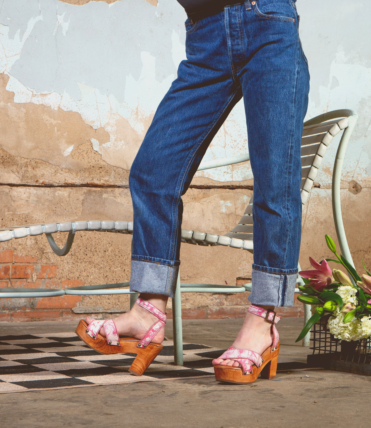 Wearing the Kalah sandals by Bed|Stü—featuring a pink platform design with an adjustable ankle strap—a person in cuffed jeans stands on a patterned tile floor near a white chair and a bouquet of flowers.