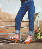 Wearing the Kalah sandals by Bed|Stü—featuring a pink platform design with an adjustable ankle strap—a person in cuffed jeans stands on a patterned tile floor near a white chair and a bouquet of flowers.