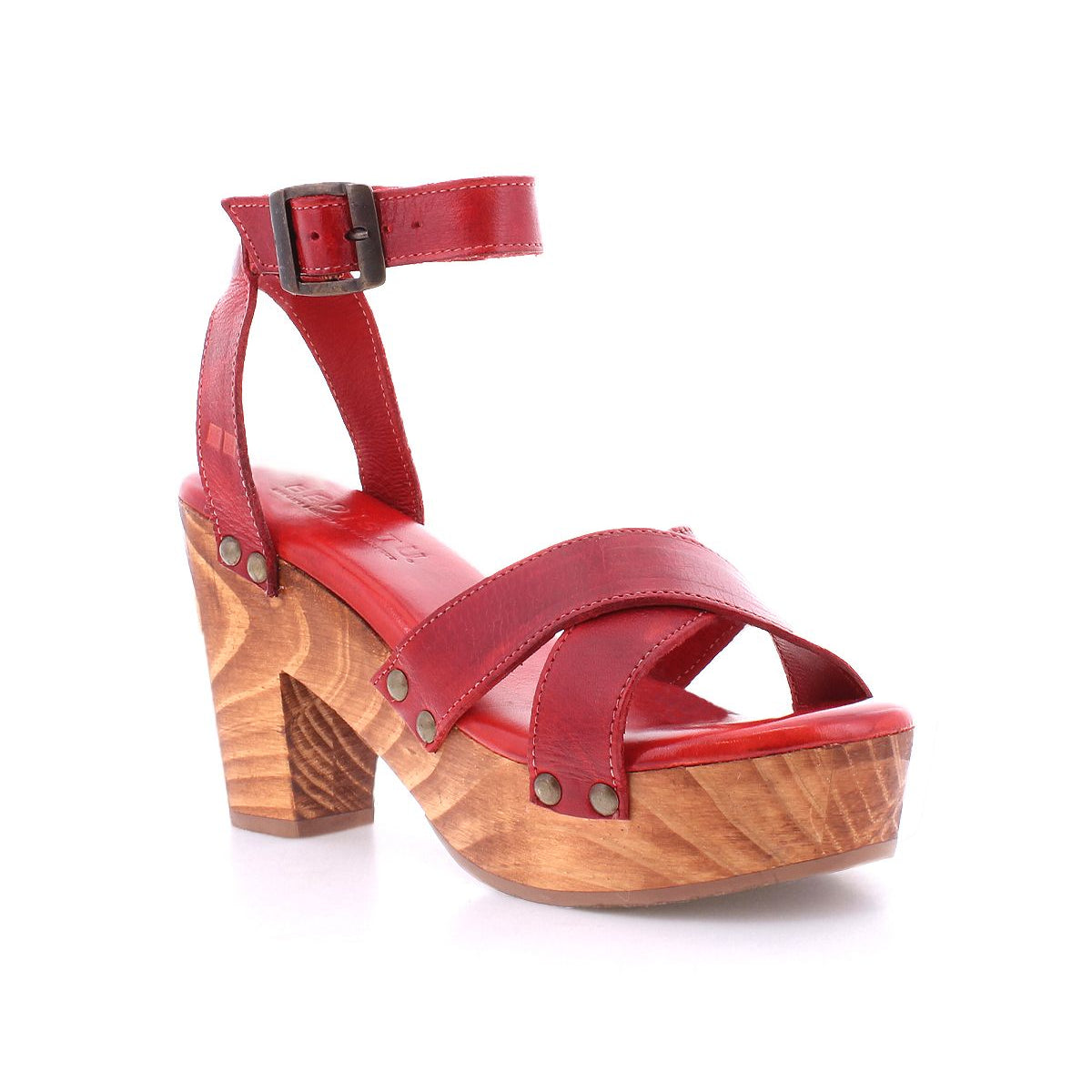 The Bed|Stü Kalah is a red leather sandal featuring an adjustable ankle strap, sustainably sourced wooden platform heel, mid-height chunky heel, and metal rivet accents, shown on a white background.