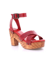 The Bed|Stü Kalah is a red leather sandal featuring an adjustable ankle strap, sustainably sourced wooden platform heel, mid-height chunky heel, and metal rivet accents, shown on a white background.