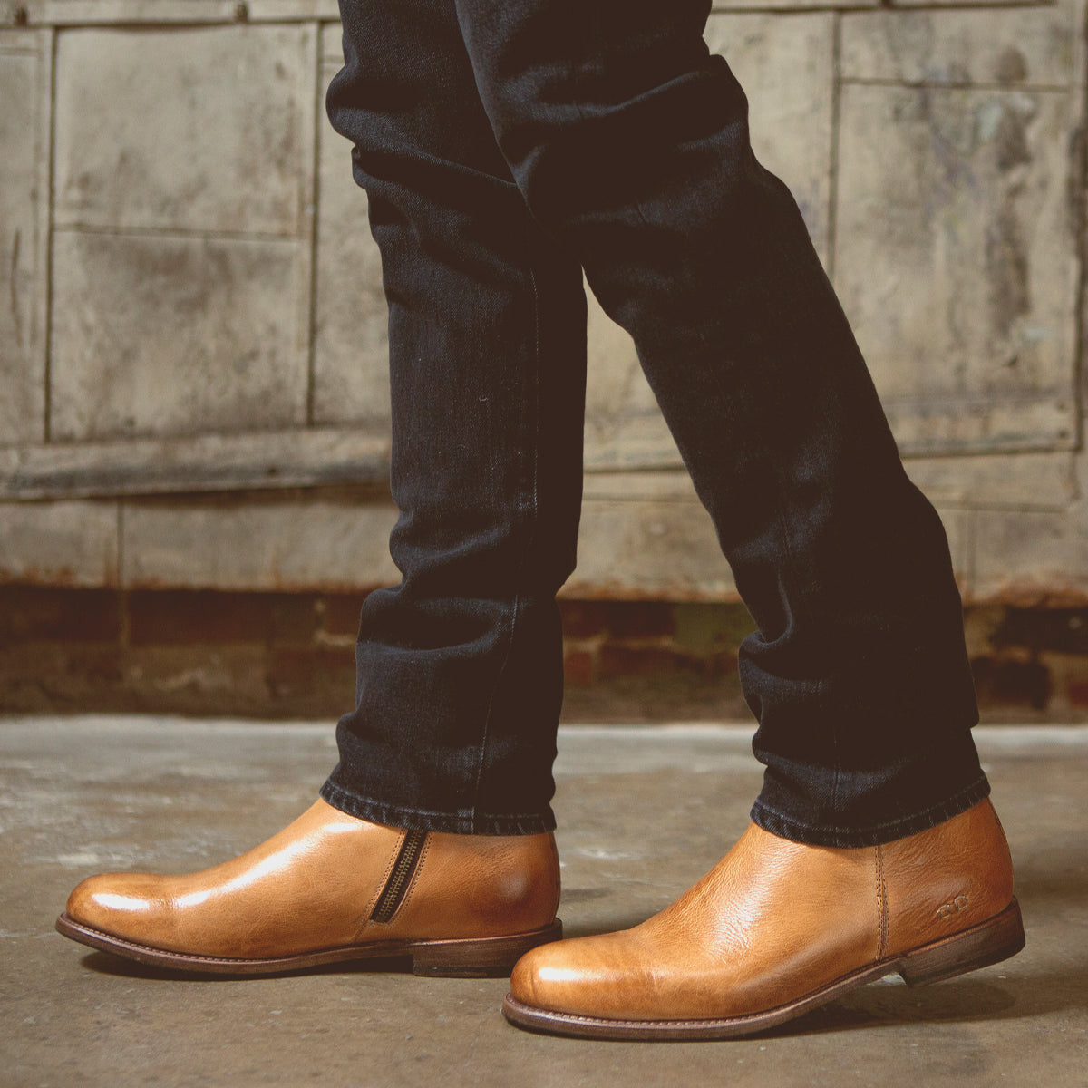 A person wearing slim-fit dark jeans and the Bed|Stü Kaldi classic tan leather ankle boots stands indoors on a concrete floor, with a worn, industrial-style wall in the background.