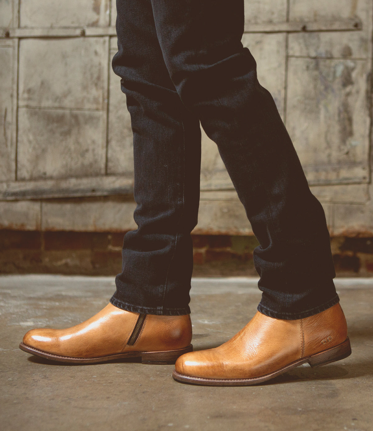 A person wearing slim-fit dark jeans and the Bed|Stü Kaldi classic tan leather ankle boots stands indoors on a concrete floor, with a worn, industrial-style wall in the background.