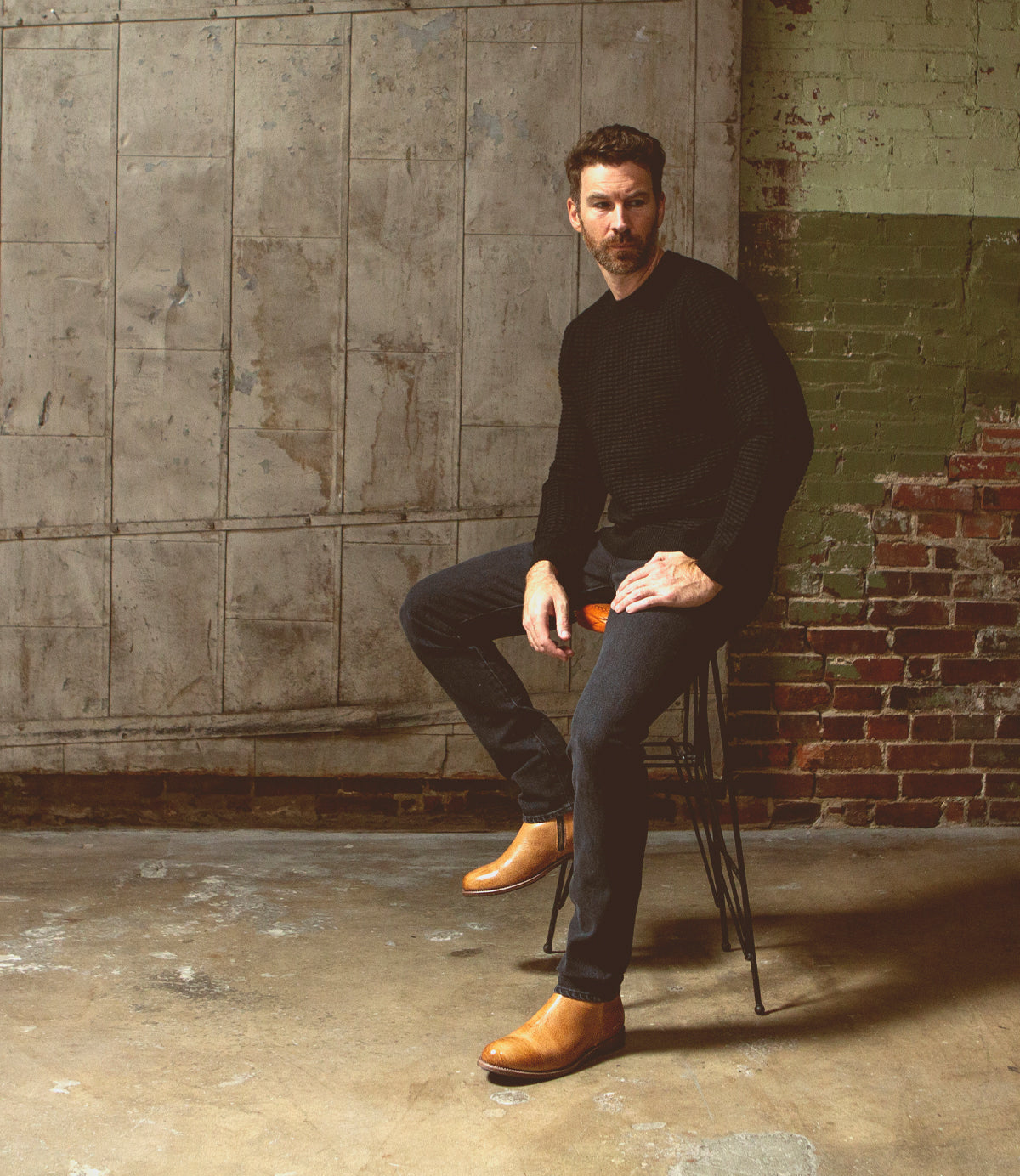A man with a beard sits on a stool against a textured wall, wearing a black sweater, dark jeans, and tan Kaldi boots by Bed|Stü.