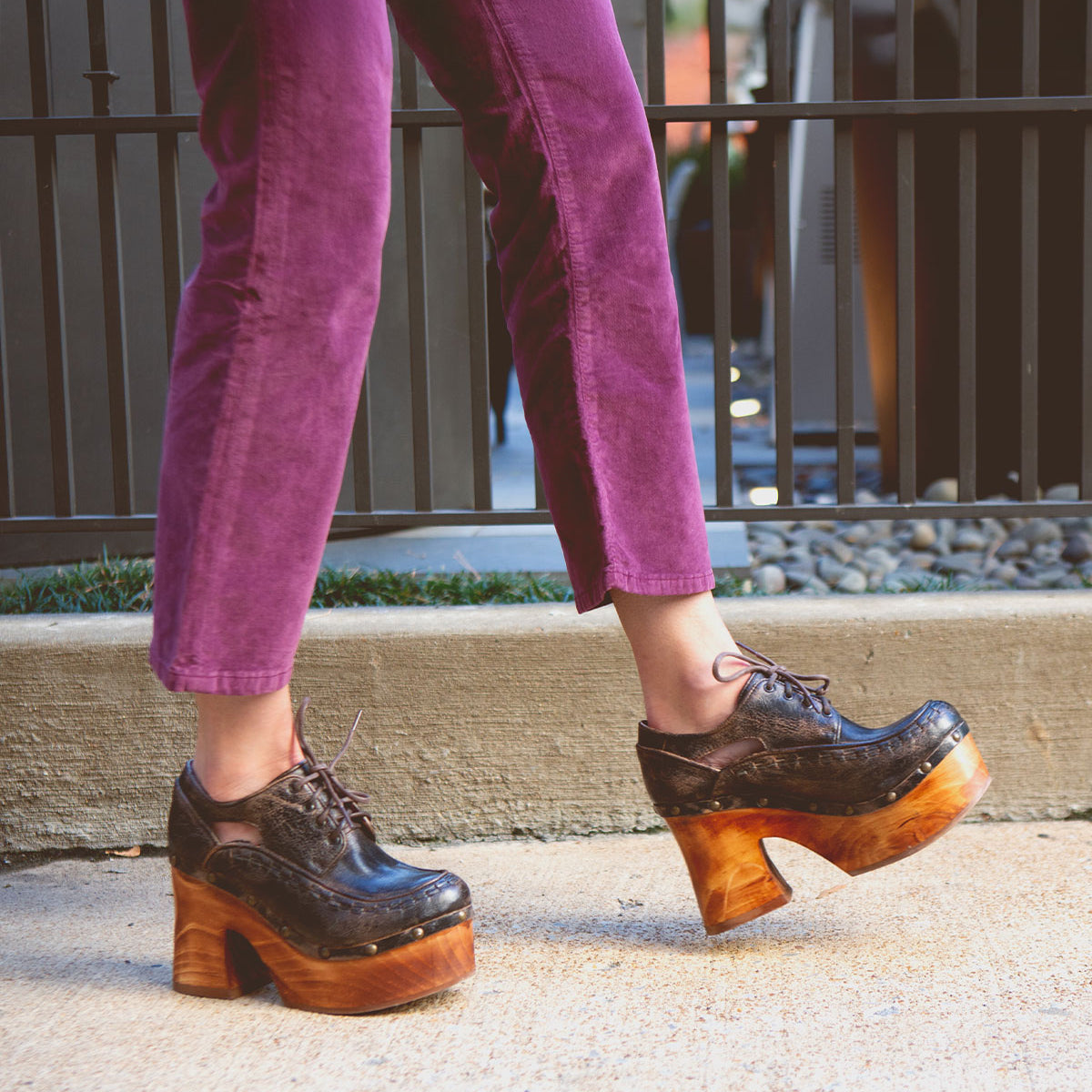 A person walks on a sidewalk wearing purple pants and the Bed|Stü Karaoke dark brown lace-up platform shoes, featuring chunky wooden heels that channel a 70s vibe.