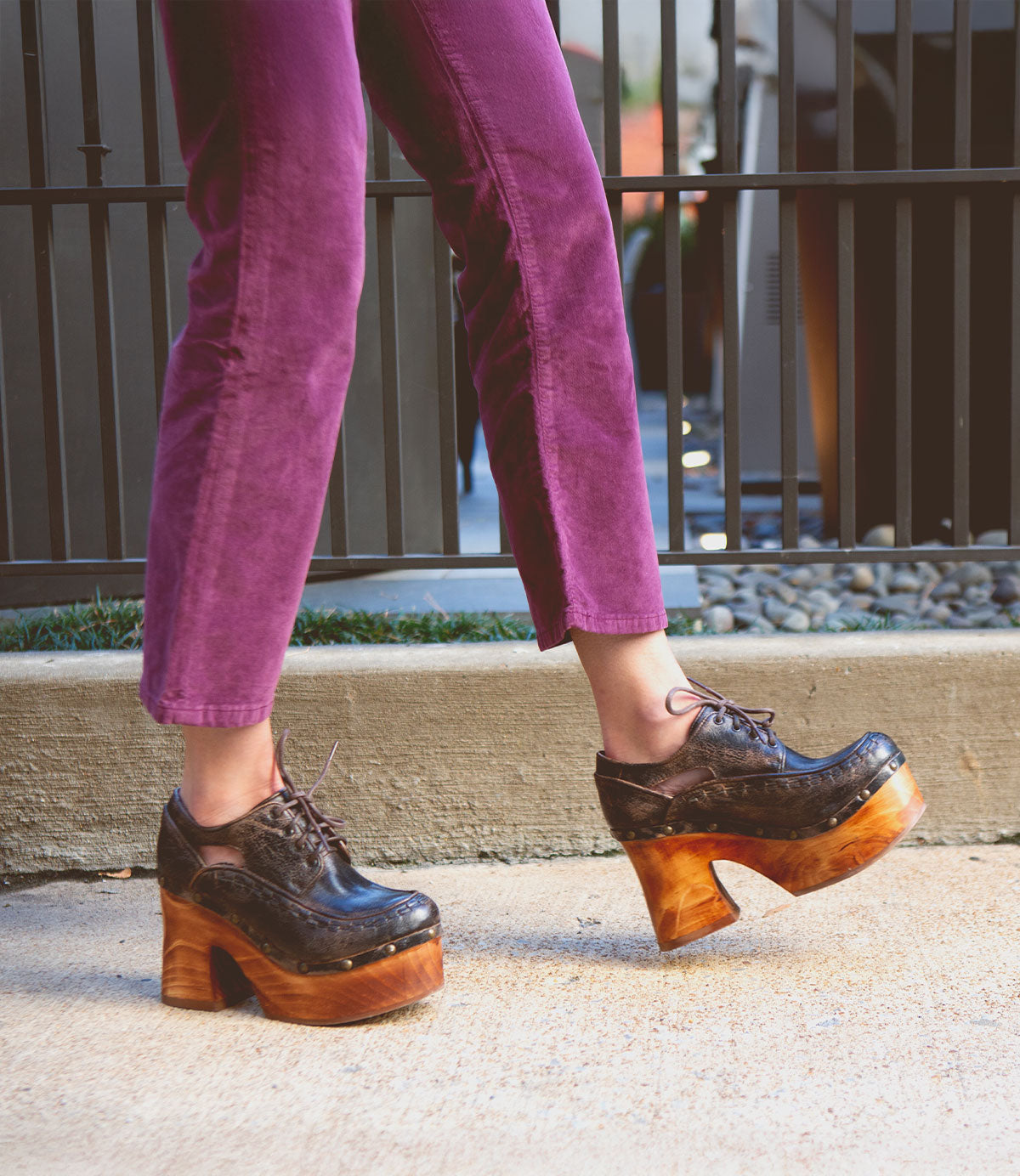 A person walks on a sidewalk wearing purple pants and the Bed|Stü Karaoke dark brown lace-up platform shoes, featuring chunky wooden heels that channel a 70s vibe.