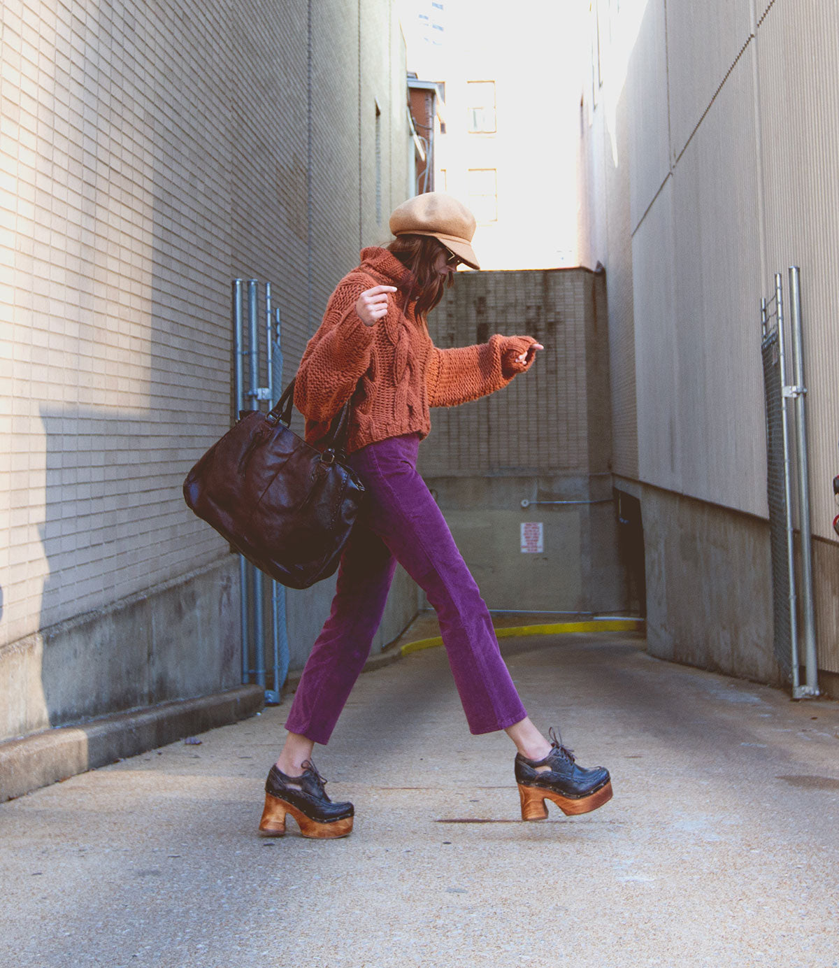 A person wearing the Karaoke chunky wooden platform shoes by Bed|Stü walks in an alley, dressed in a brown hat, orange sweater, purple pants, and carrying a large dark bag.