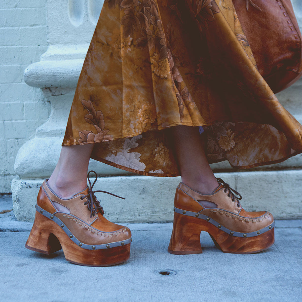 A woman standing on a sidewalk wears a long brown floral dress paired with Bed|Stü's Karaoke lace-up chunky wooden platform shoes featuring cut-out heels.