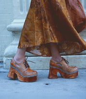 A woman standing on a sidewalk wears a long brown floral dress paired with Bed|Stü's Karaoke lace-up chunky wooden platform shoes featuring cut-out heels.